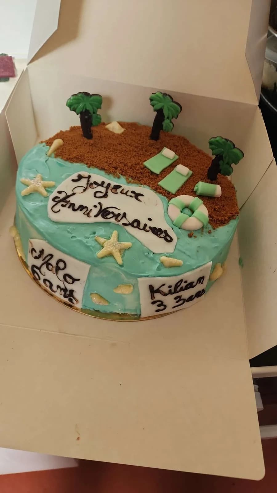 Gâteau turquoise d’O’délices d’Élodie, décoré de palmiers et de transats, dans une boîte à gâteau ouverte.
