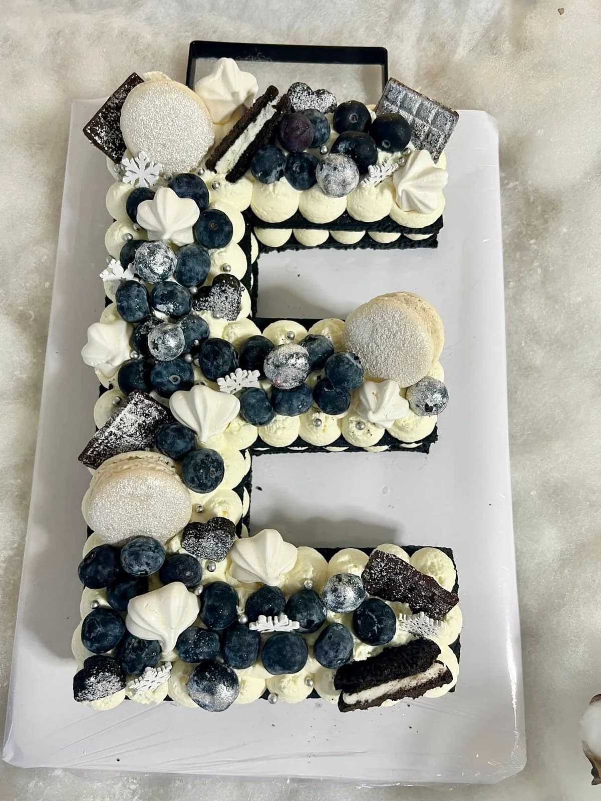 Letter cake en forme de E d’O’délices d’Élodie, garni de crème, de myrtilles, de macarons et de biscuits.