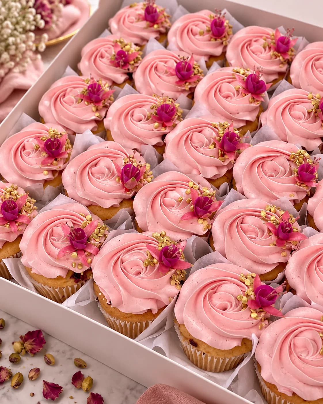 Cupcakes roses d’O’délices d’Élodie dans une boîte mise en scène avec des fleurs et du linge rose.